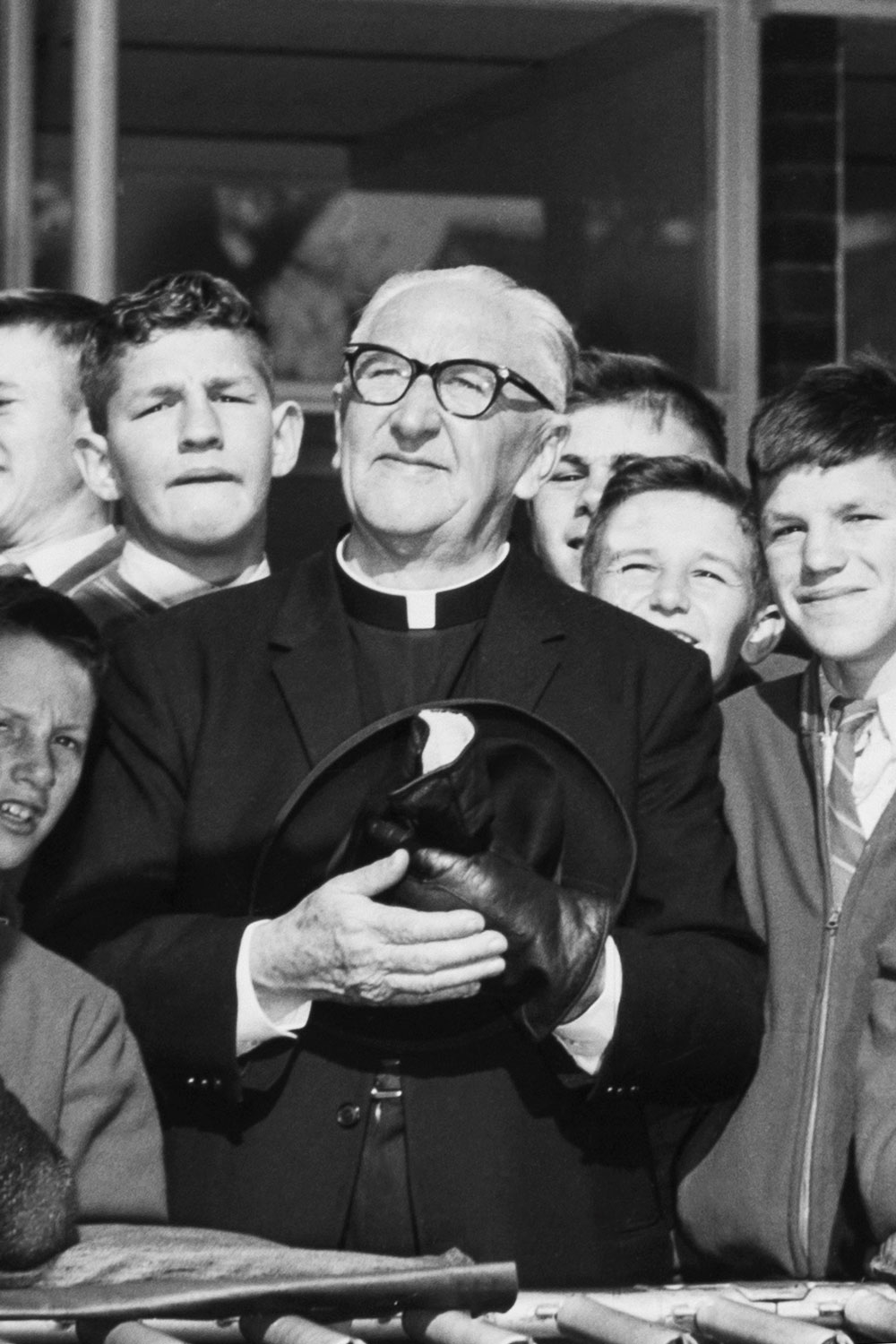 Image 2 of 5 - A closeup of Father Dunlea surrounded by a group of smiling boys at Boys Town in Engadine, now known as the Dunlea Centre. Father Dunlea holds his hat and gloves in his hands in a moment of reflection.