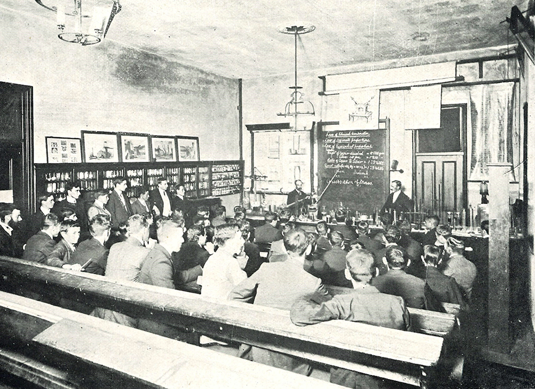 Black and white image of students sitting down in a classroom facing the blackboard, backs towards the camera. Two men standing at the front of the room on either side of the blackboard.