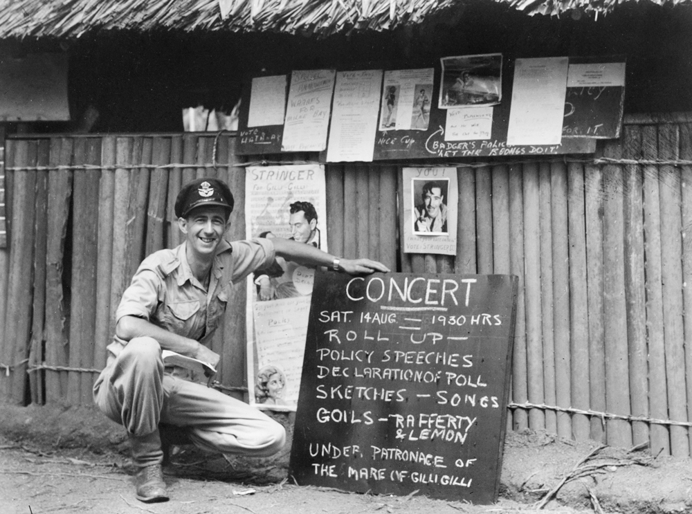 Image 2 of 4 - Black and white image of Chips Rafferty crouching down next to a concert sign. Chips is wearing boots, pants, a shirt and a military hat. Behind the main concert sign is some more posters and papers on a fence.