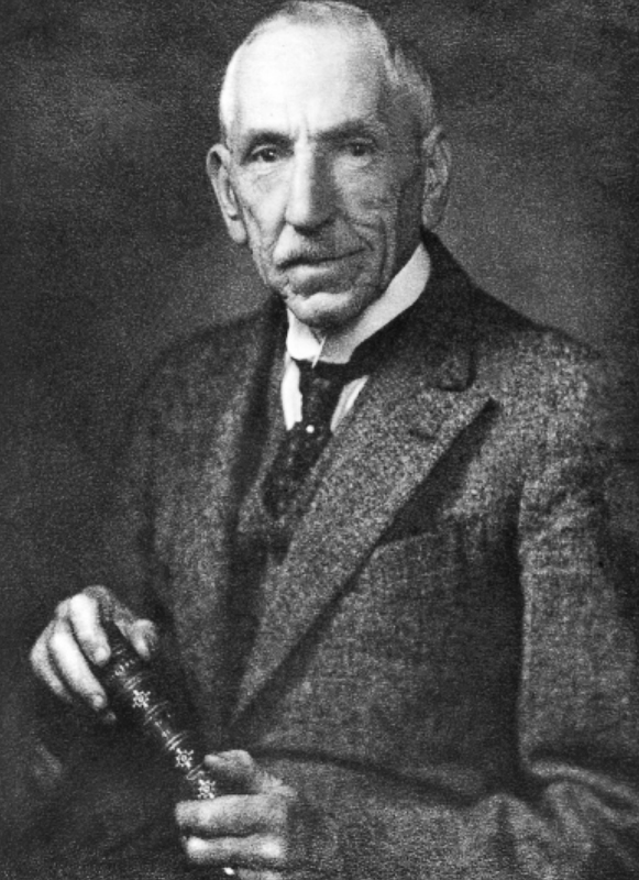 Black and white portrait of Billy Hughes wearing a suit and tie with a book in-hand.