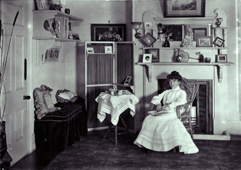 Image 5 of 5 - Black and white image of Ethel Turner sitting on a chair in her home. Behind her is a fireplace and the room contains décor, furniture and little images on shelves.
