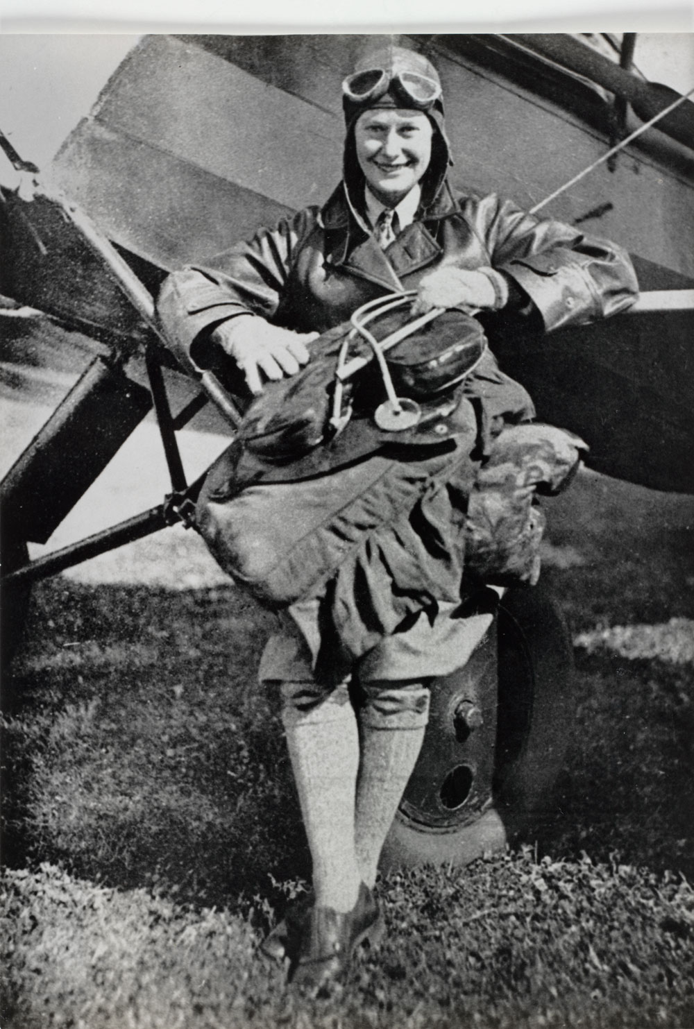 Image 2 of 5 - Black-and-white photo showing Nancy Bird Walton standing casually in front of an old-fashioned aircraft wearing full aviation gear, including a cap, with goggles pushed up over her forehead