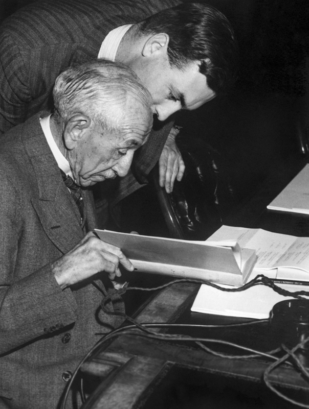 Image 4 of 6 - Black and white image of Billy Hughes sitting down accompanied by another man looking over his shoulder, both viewing a document.