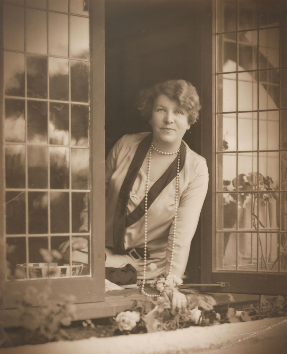 Black and white portrait of Ethel Turner sitting on a windowsill looking outside. There are flowers just outside the windowsill and she is wearing a long sleeve top and a long pearl necklace.
