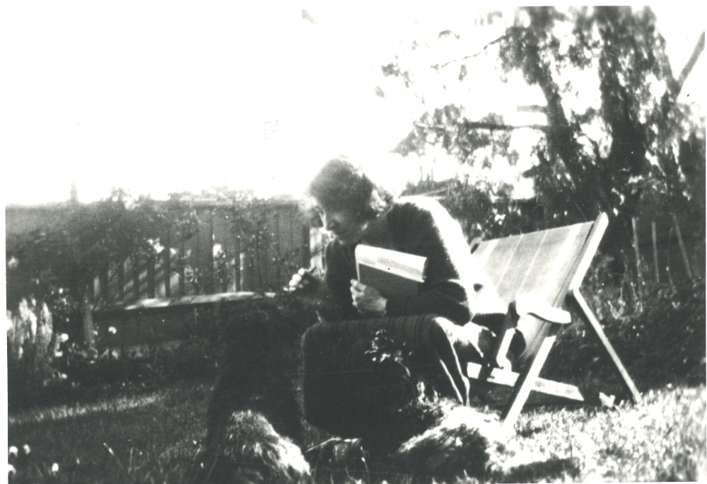 Image 4 of 5 - Black and white image of May Gibbs sitting outside with her Scotch Terriers. May is holding a book close to her chest with her left hand as she leans forward in the chair.
