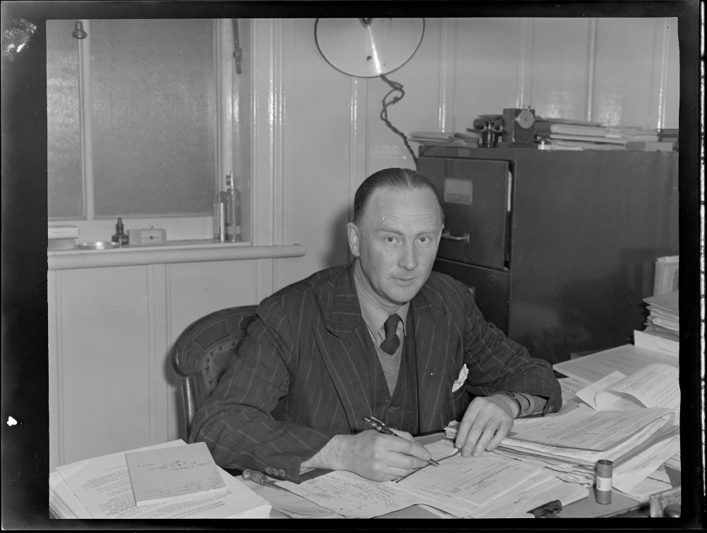 Image 4 of 5 - Black and white portrait of a man in a suit sitting down at a desk with a pen in hand.