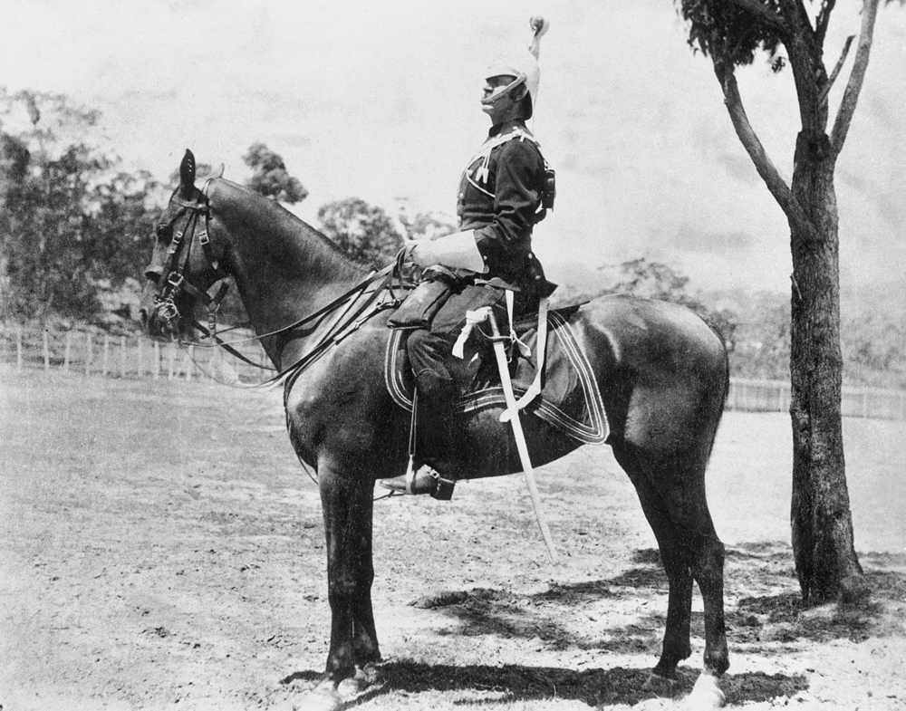 Image 5 of 7 - Black and white image of a man sitting on a horse. The man is wearing an army uniform and has a sword hanging off his left leg. There is a tree next to them on the right and forestry in the background.