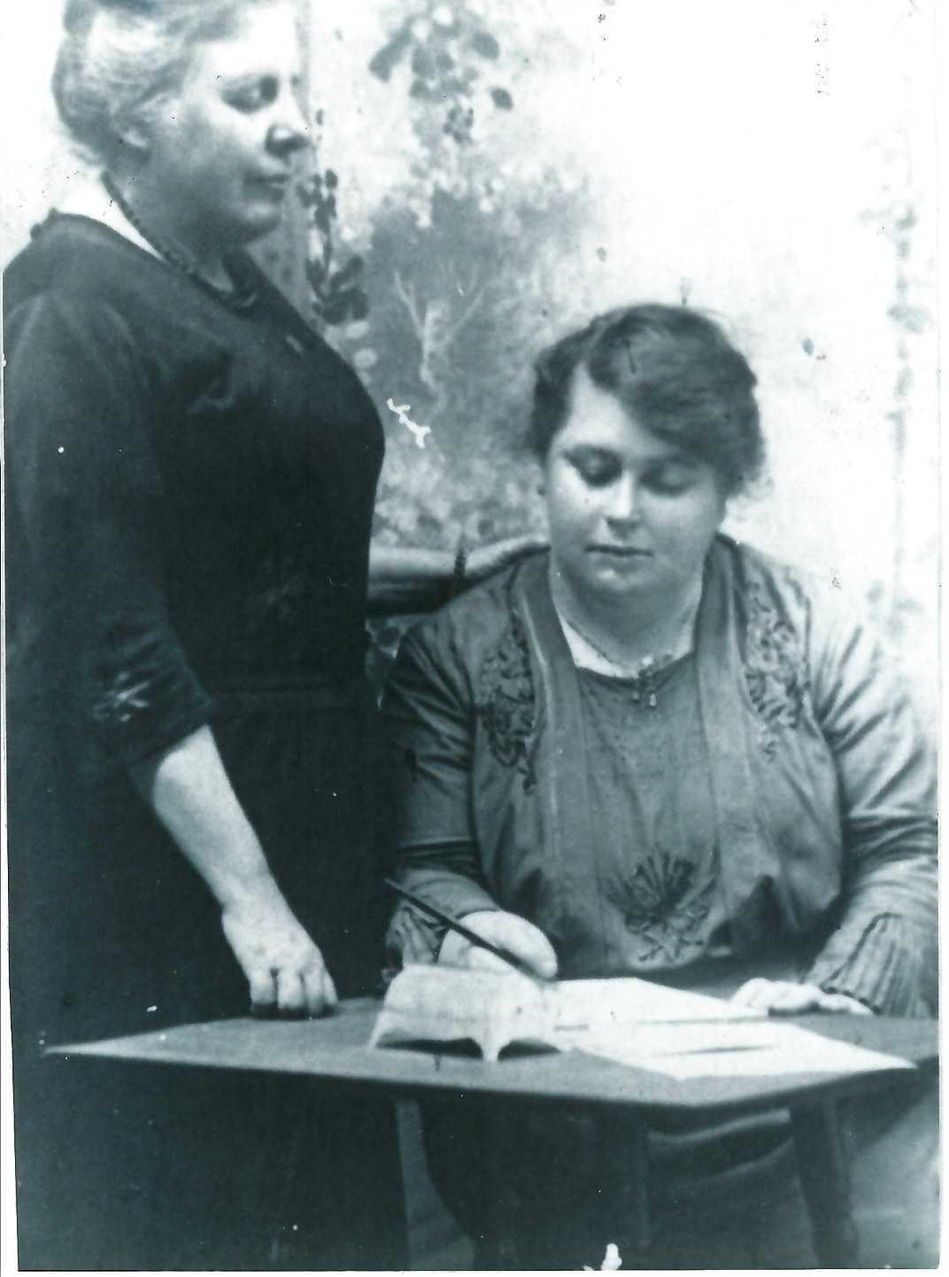 Image 1 of 4 - Black and white photograph of Susan, standing with her hand on the shoulder of a female seated pupil.
