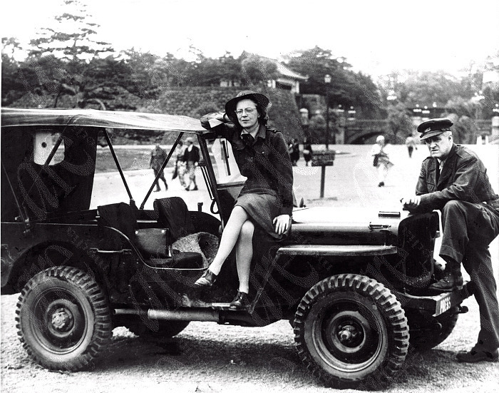 Black and white photograph of a woman sitting on a car, while a man leans his leg against the front of the car