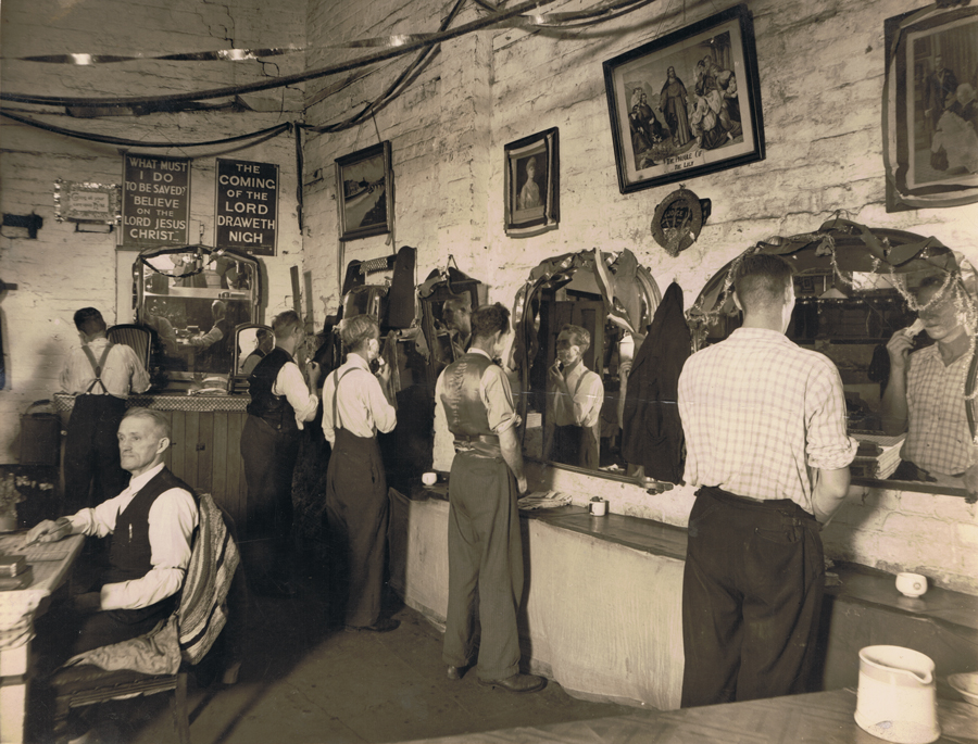 Image 3 of 4 - Black and white image of 5 men shaving their faces in front of mirrors. The room is decorated with pictures and signs above the mirrors. Arthur Stace is sitting down at a desk on the left-hand side of the image. All the men are wearing pants and button up shirts.