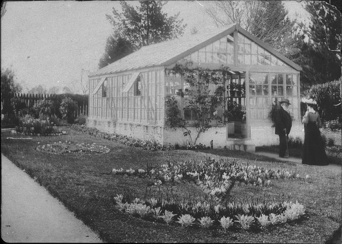 Image 1 of 4 - Black and White image of a garden with grass and plants in the foreground and a white greenhouse in the background. In front of the greenhouse is Daniel Gowing and a woman.