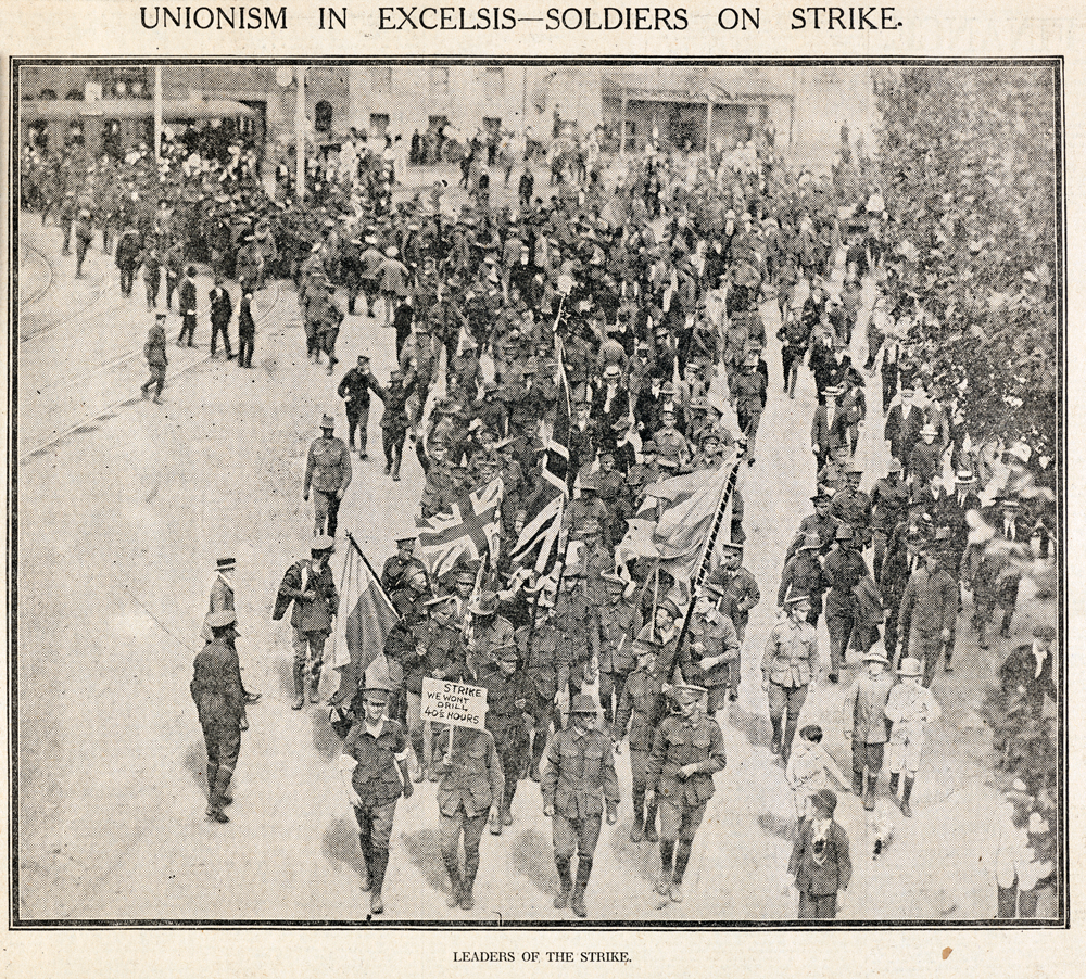 Image 5 of 6 - Black and white image taken from a newspaper of hundreds of soldiers marching down the street with the leaders in the front holding the flags. Above the image says, ‘UNIONISM IN EXCELSIS – SOLDIERS ON STRIKE’ and below the image ‘LEADERS OF THE STRIKE’.
