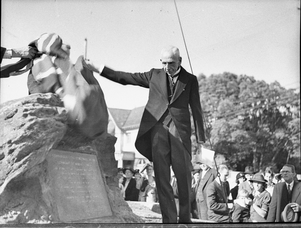 Image 5 of 6 - Black and white image of a Billy Hughes outside unveiling a plaque on a rock. Billy Hughes is wearing a suit and there are people behind him.