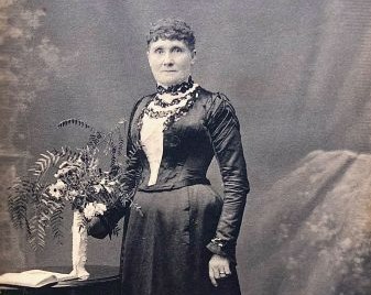 A formal portrait of Bessie Robinson wearing a dark dress, standing next to a table with a vase of flowers. 
