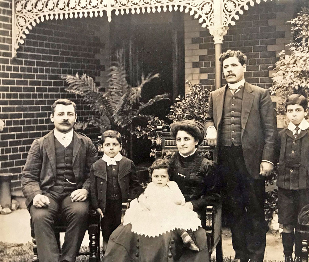 A portrait of Betro Abicare and family standing in front of a house.    The man sitting at the left is believed to be his brother Nicholas, continuing L to R Victor, baby Frederick, Julia (seated, holding Frederick), Betro (standing) and Claude 