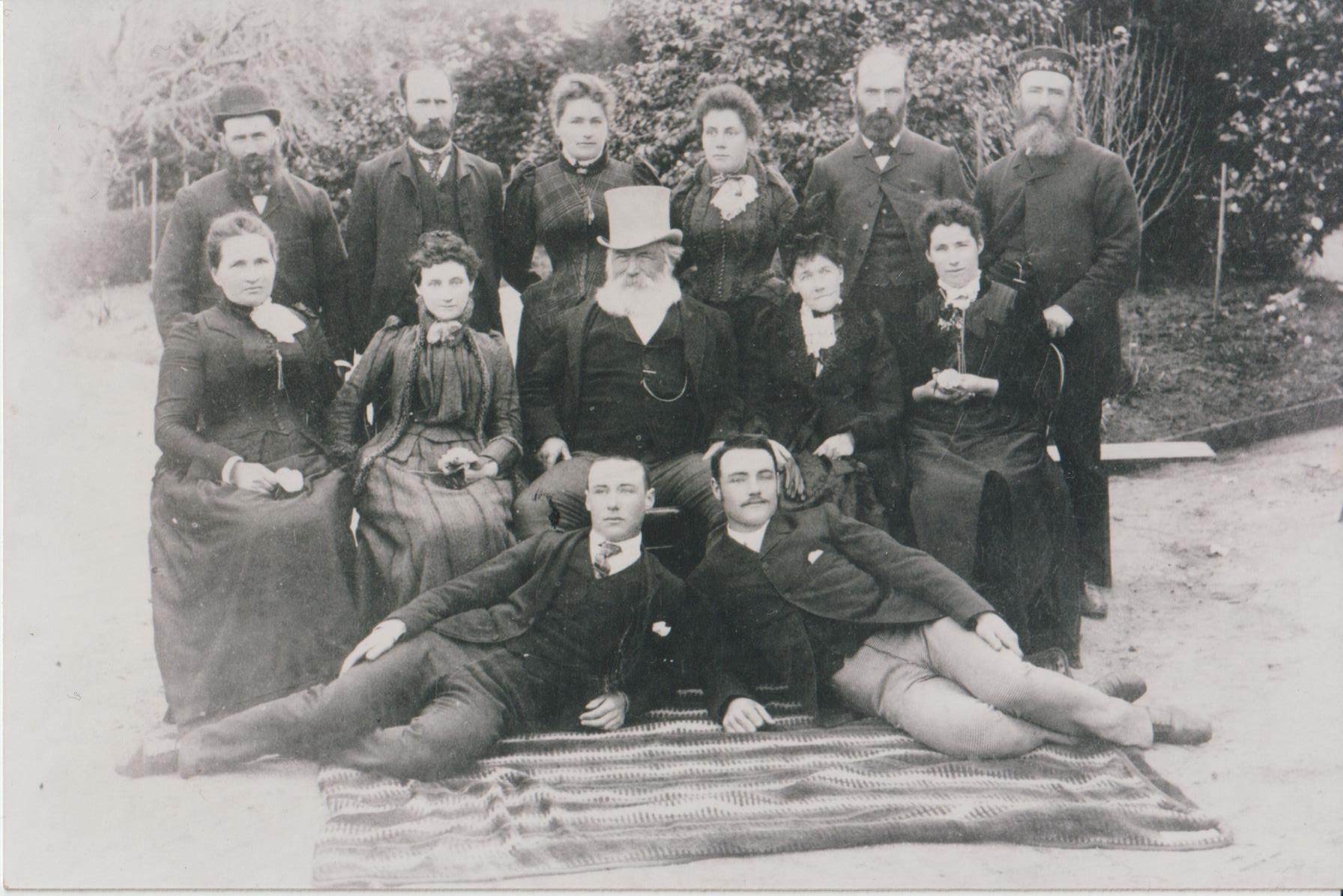 Image 3 of 4 - Black and white image of 7 men and 7 women. The men are wearing suits, with three of them wearing hats and the women are wearing Edwardian dresses. Two of the men are lying down in front of the others who are sitting and standing behind them. The family is standing outside with bushes and trees behind them.