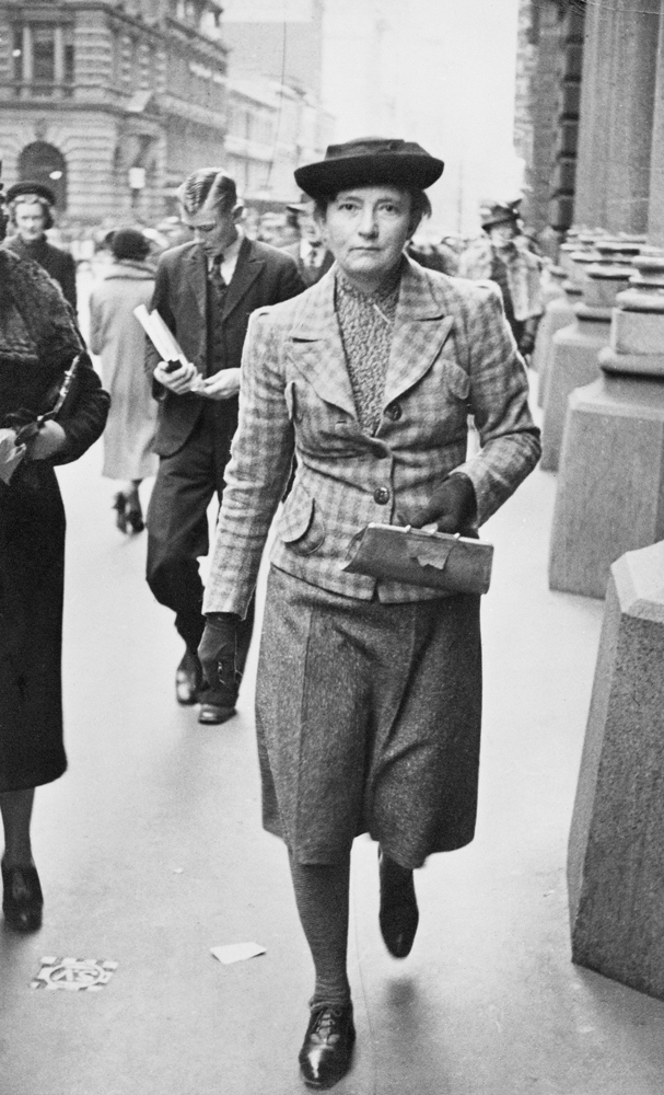 Image 2 of 6 - Black and white image of Grace Cossington Smith walking down the street with other people walking in the background. She is holding a small purse while wearing shoes, stockings, a skirt, a plaid blazer, gloves and a hat.