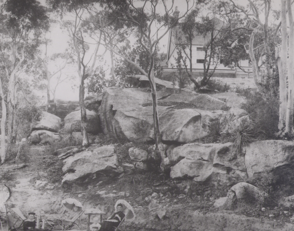 Image 4 of 7 - Black and white image of a family picnicking in the foreground with trees and large rocks behind them. On top of the rocks is a house.