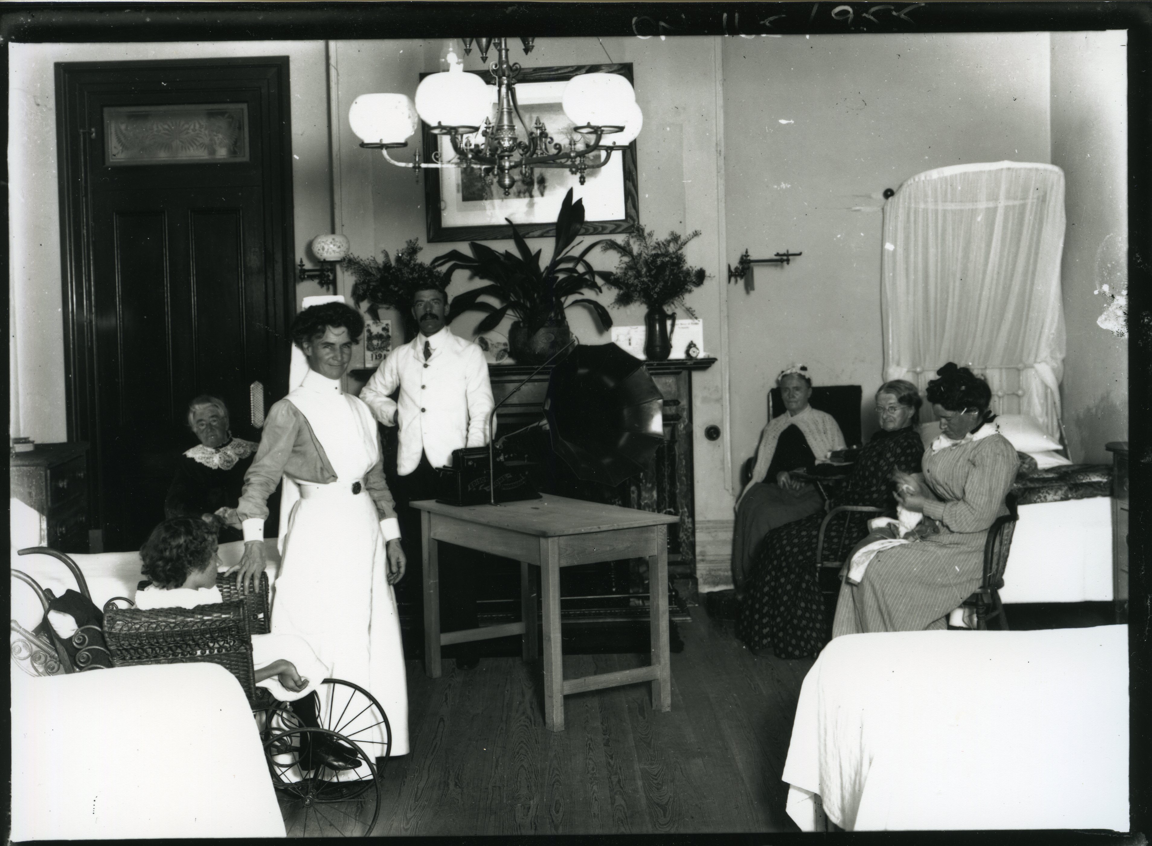 Image 3 of 4 - Black and white photo of a room. Standing in the room is a woman wearing a white nurse’s outfit and a man wearing a white suit. Patients are sitting on a bed nearby. In the middle of the room is a gramophone.