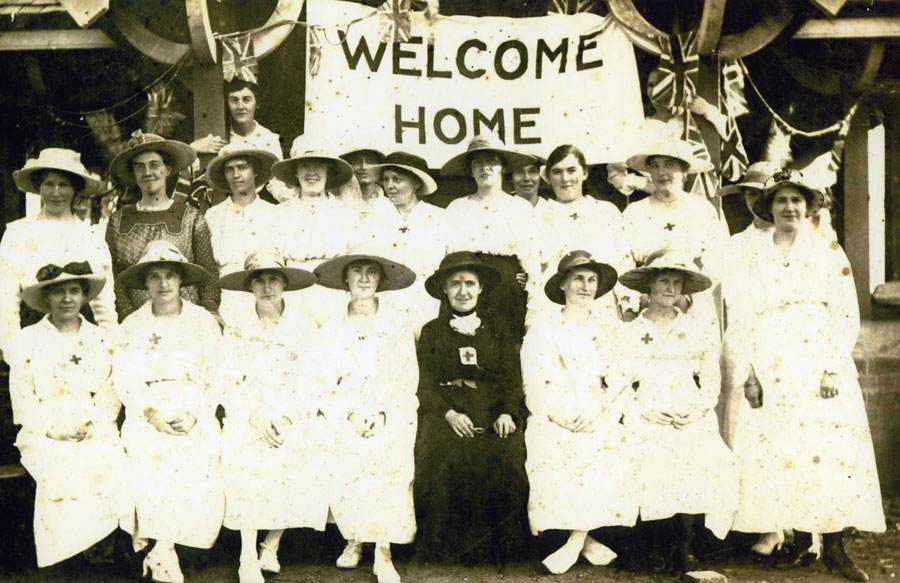 Image 2 of 5 - Photograph of the ladies of Camden Red Cross Sewing Circle taken in Macarthur Park standing under a Welcome Home banner. Woman in black is Mrs Honor Harris (nee Sidman)