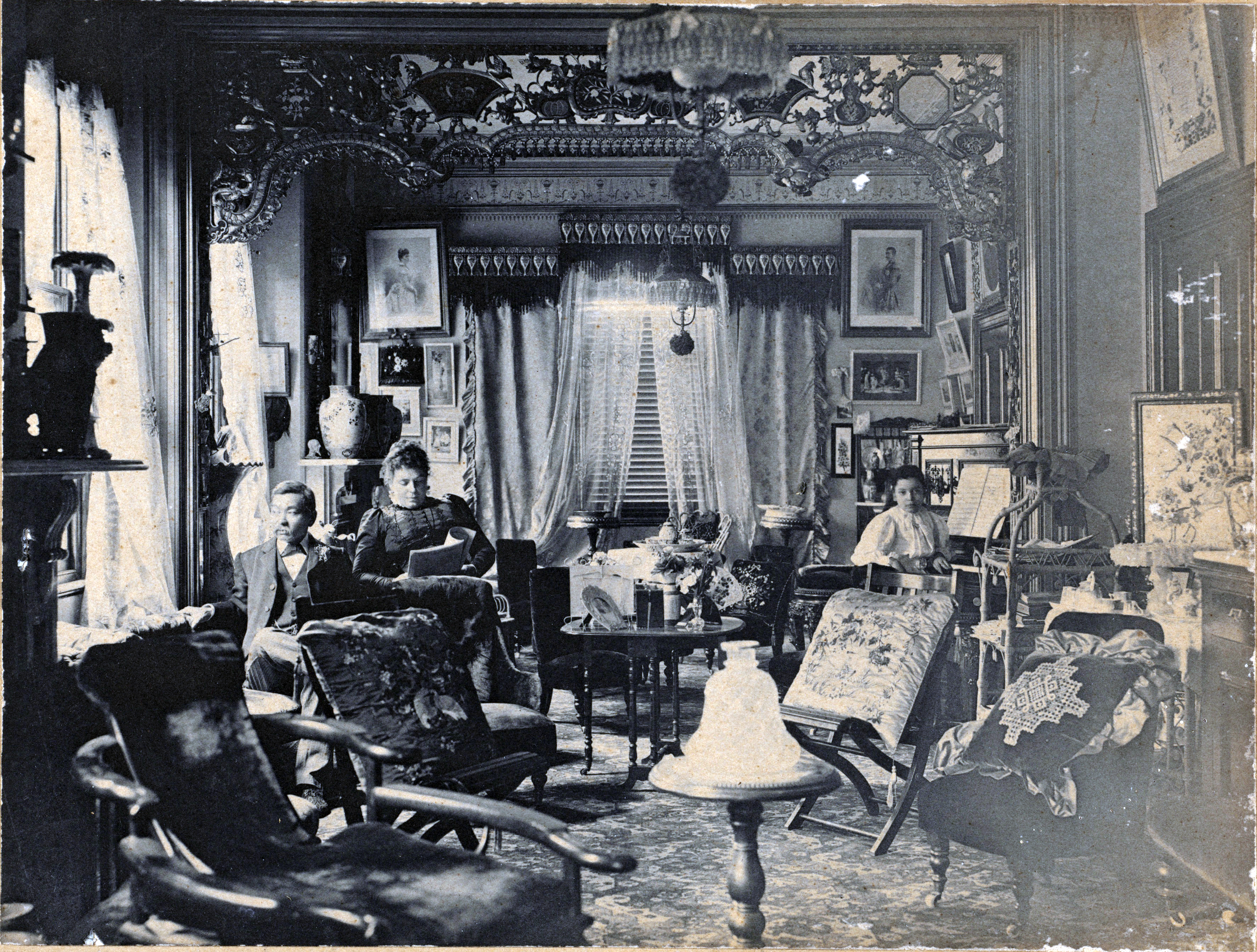 Image 2 of 6 - Black and white image of a room with wooden chairs, paintings and décor. Quong sitting on the left-hand side, looking out a window with his wife next to him reading a magazine. One child sitting to the right of the image looking at the camera.