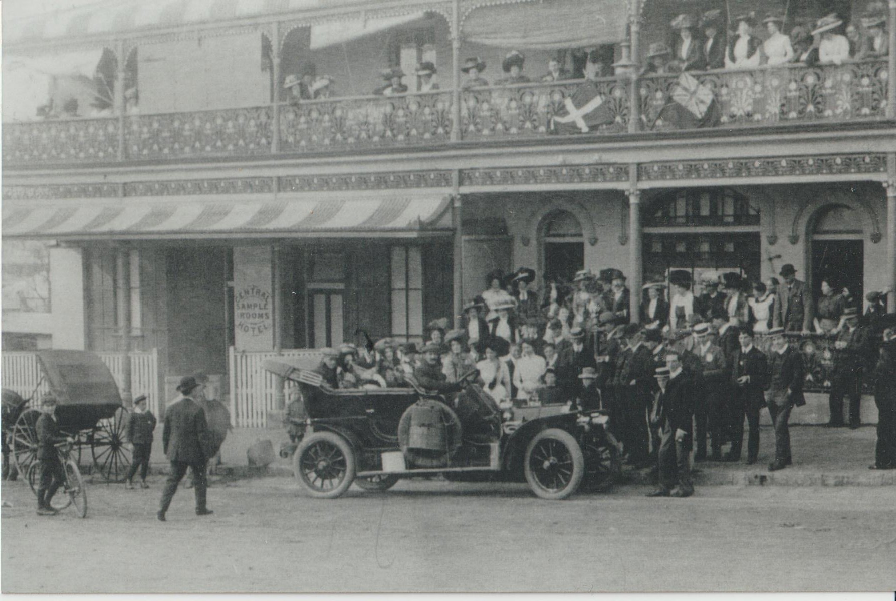 Image 2 of 4 - Black and white image of men and women in an old motor car. The car is on the street next to a building which is a hotel, with people looking out from the bottom floor entrance and on the balcony above.