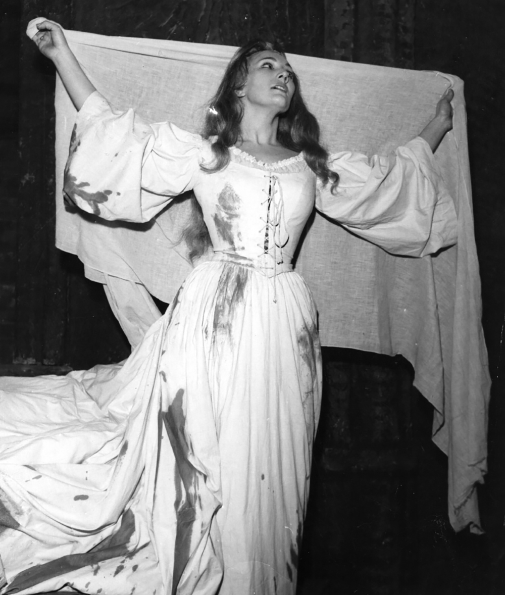 Image 4 of 5 - Black and white image of June Bronhill performing. June is wearing a white dress with long puff sleeves. Her arms are out as she holds a white linen cloth.