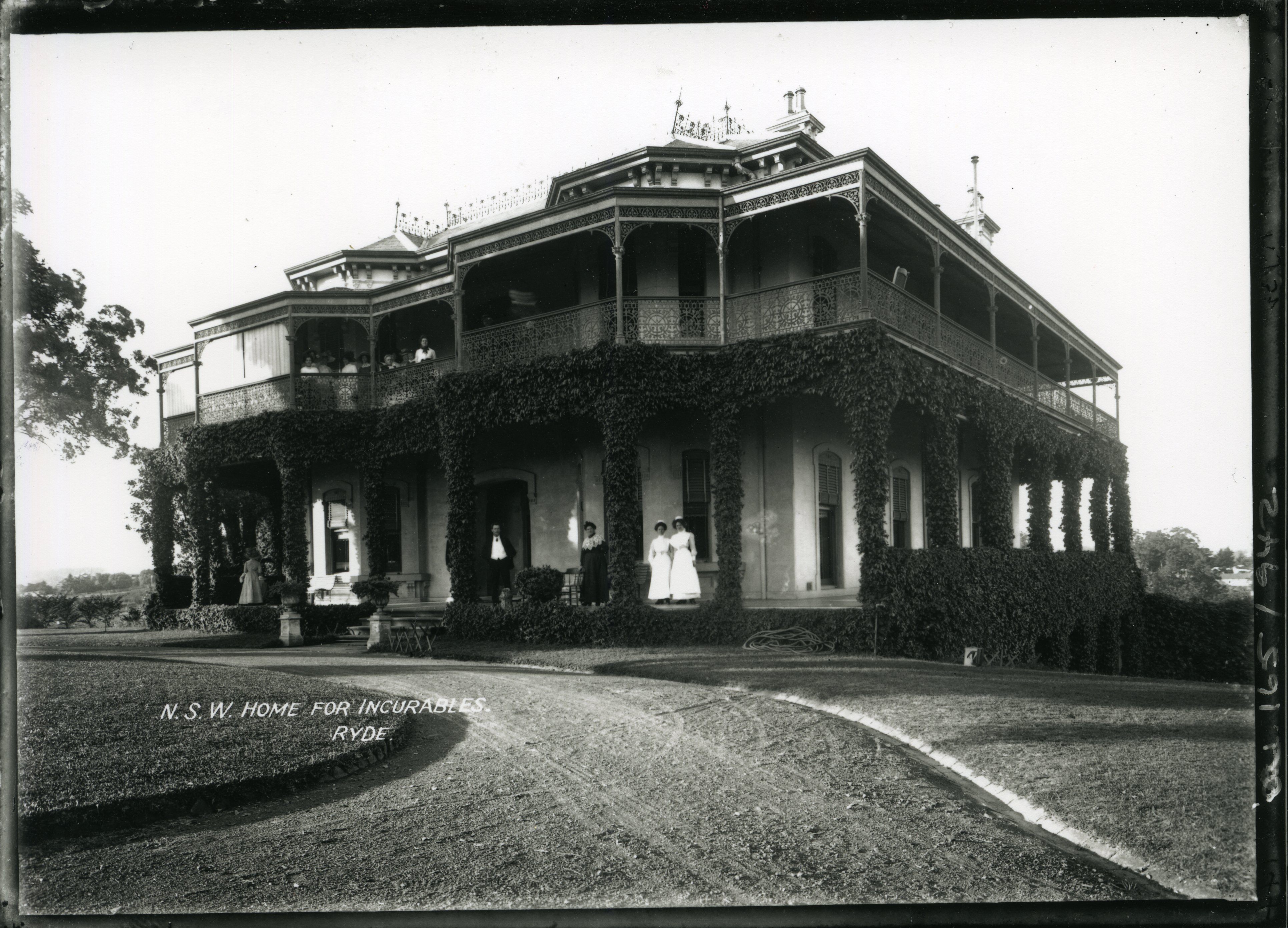 Image 4 of 4 - Black and white photo of a large two-story house with a wraparound balcony and veranda. Standing on the veranda and balcony are nurses and patients.