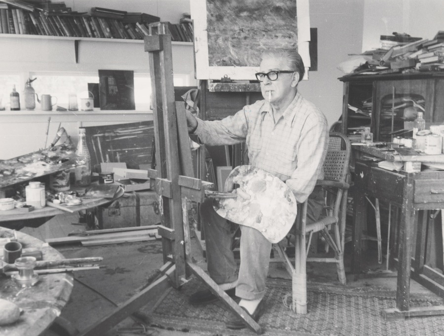A black and white photo of a man wearing glasses who is painting at an easel while sitting down on a wooden chair  
