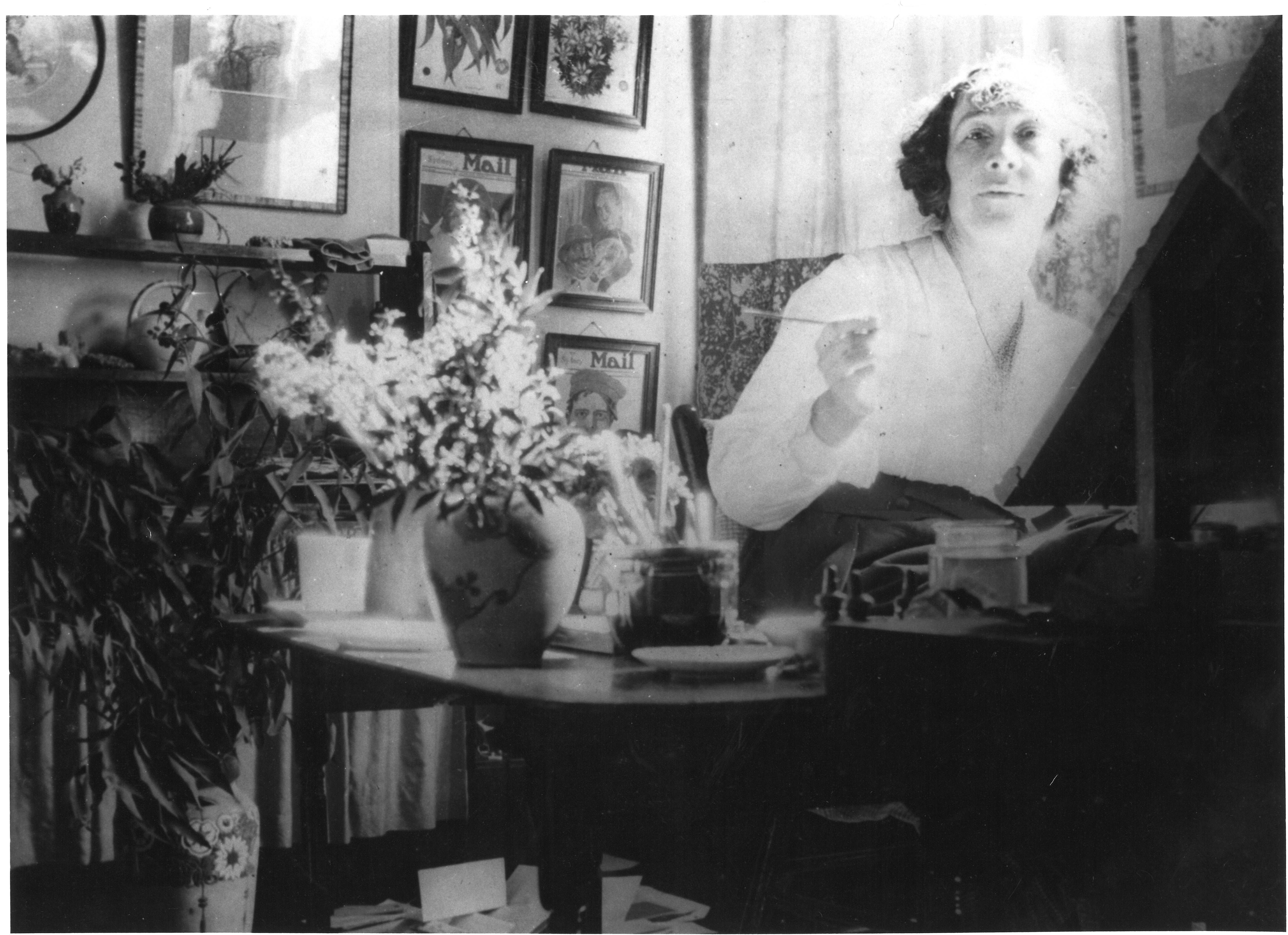 Photograph of May Gibbs working at her rented studio on Bridge St in Sydney. May is seated behind an easel, looking to camera holding a paintbrush. On a table next to her is a vase of native foliage and a series of her artworks for the Daily Mail are framed on the wall behind her. 