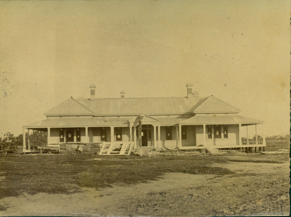 A photograph of the front façade of Bishop’s Lodge during its construction between 1889 and 1890. Pallets of construction material lie in front of the house and a workman stands atop a ladder. 