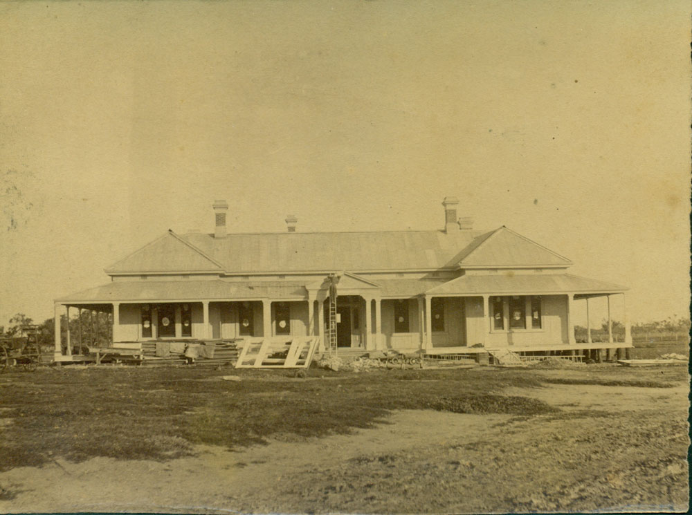 Image 3 of 6 - A photograph of the front façade of Bishop’s Lodge during its construction between 1889 and 1890. Pallets of construction material lie in front of the house and a workman stands atop a ladder.