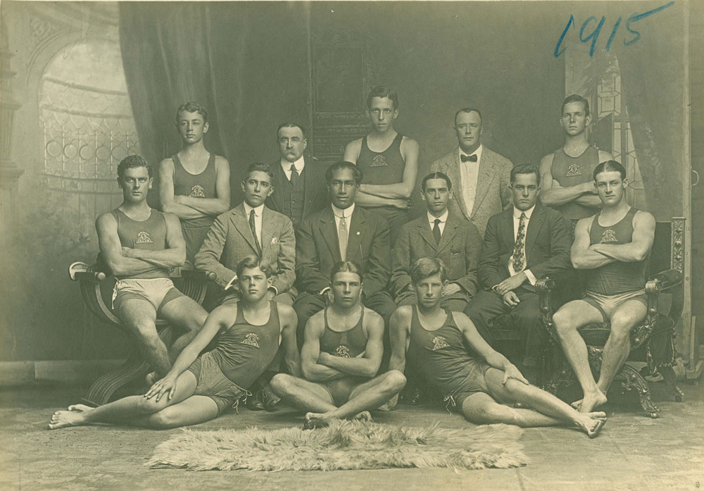 Formally posed portrait of Duke Kahanamoku, seated centre wearing a suit, surrounded by Manly Surf Life Saving Club Members taken in 1915 