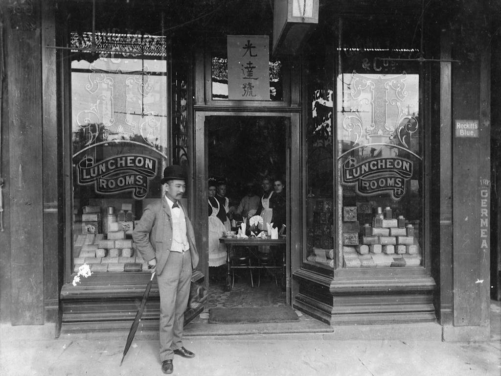 Black and white image of Quong Tart standing in front of a store front. Entrance is open with people standing inside, gazing at the camera. Windows on the left and right with the text ‘Luncheon Rooms’, and Chinese text on a window above the door frame.