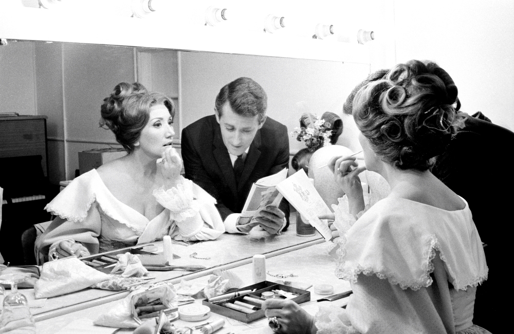 Image 5 of 5 - Black and white image of June Bronhill applying her make-up in front of the dressing room mirror with her manager. June is wearing a dress and the man is wearing a suit.