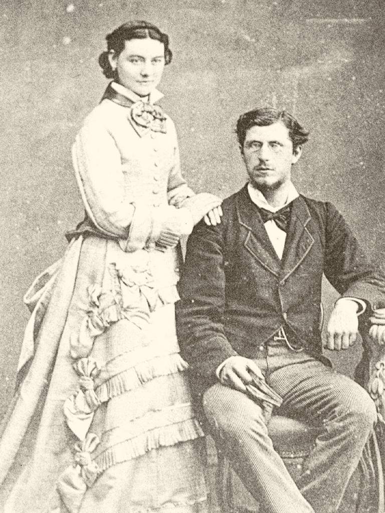 Black and white portrait of Thomas Fiaschi sitting down and Kate Fiaschi standing next to him leaning on his right shoulder. Thomas is wearing a suit and bowtie while Kate is wearing a dress and cravat (necktie).