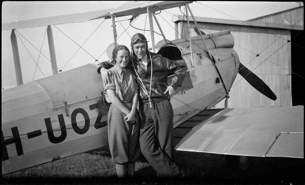 Image 5 of 5 - Black and white image of a man and a woman standing in front of an old aeroplane with ‘H-UOZ’ on the side.
