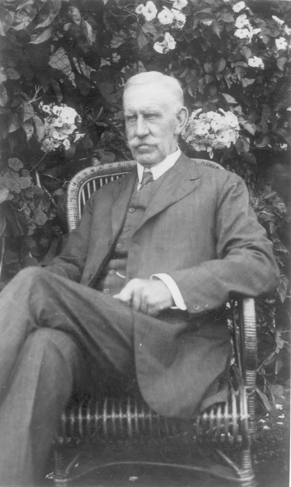 Image 3 of 7 - Black and white portrait of a man in a suit sitting on a chair outside with leaves and flowers behind him.