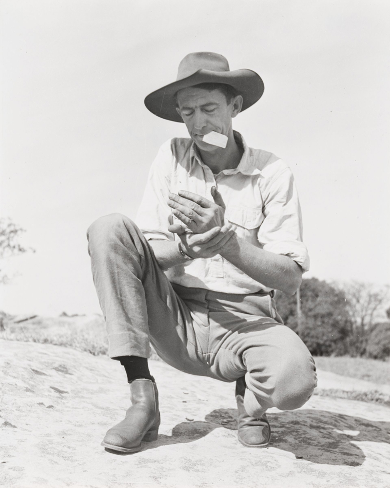 Black and white portrait of Chips Rafferty crouching down outside, wearing boots, pants, a shirt and a wide brimmed hat.