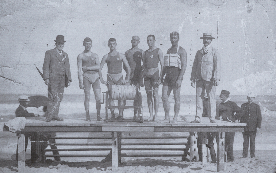 Image 2 of 3 - A black and white photo of a group of men posing for a photo on a platform on the beach. In front of them is a surf lifesaving reel