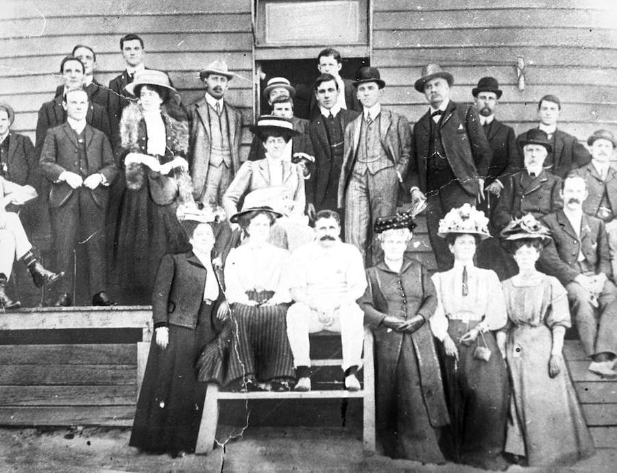 A black and white photo of a group of people in Edwardian clothes posing for a photo in front of a weatherboard shack 