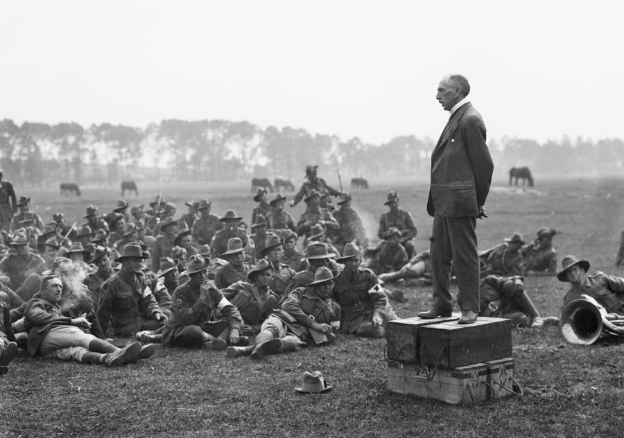 Image 2 of 6 - Billy Hughes standing on four ammunition boxes, addressing men a part of the 5th Australian Field Ambulance on an open grass field with horses in the background.