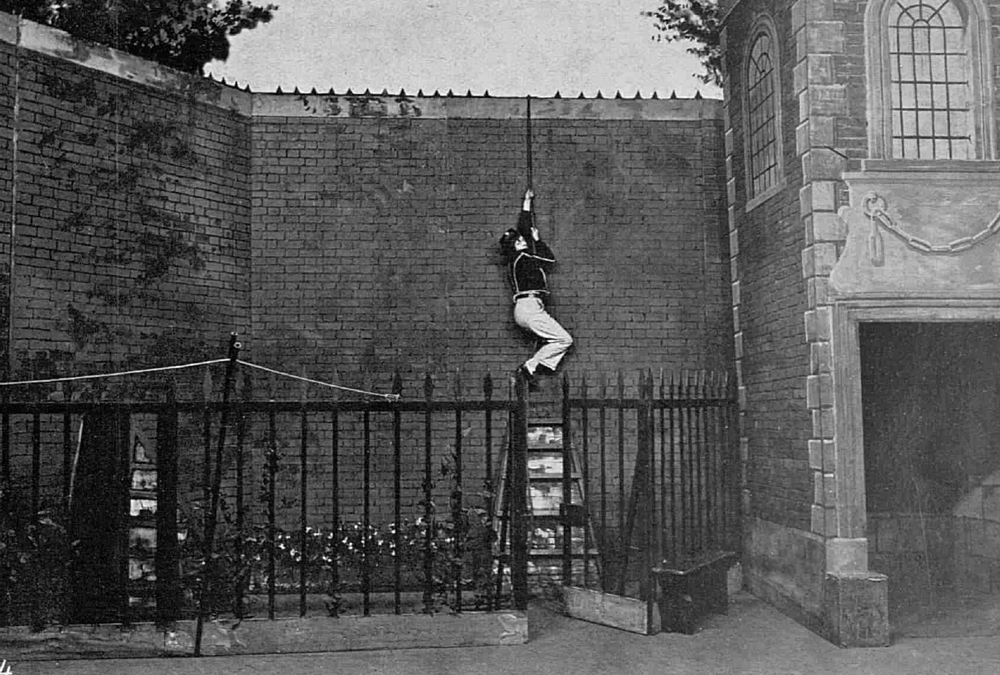 Image 2 of 6 - A woman dressed as a sailor holding onto a rope, climbing up a tall brick wall. A small ladder is underneath her, surrounded by small plants and a fence.
