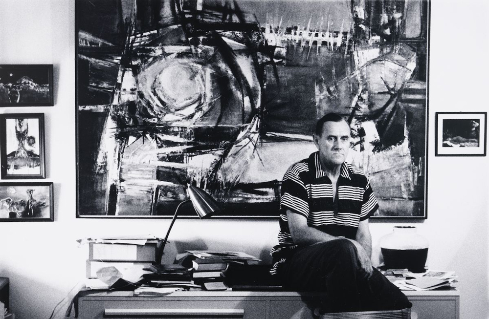 Image 3 of 6 - Black and white image of Patrick White sitting on his desk. There are pictures framed on the wall behind him and a clutter of items on the desk. Patrick is wearing a striped short-sleeve polo and pants.