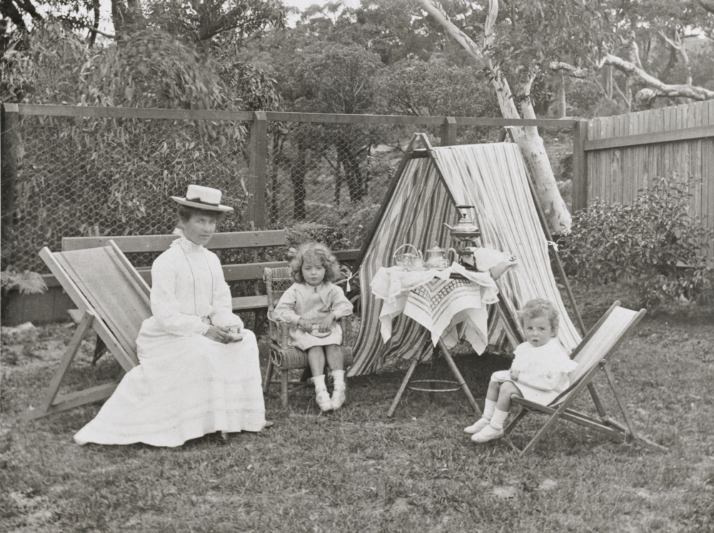 Image 4 of 5 - Black and white image of Ethel Turner and her children outside having a tea party. Ethel is to the left, wearing a dress and a hat. In the background is a fence and trees.