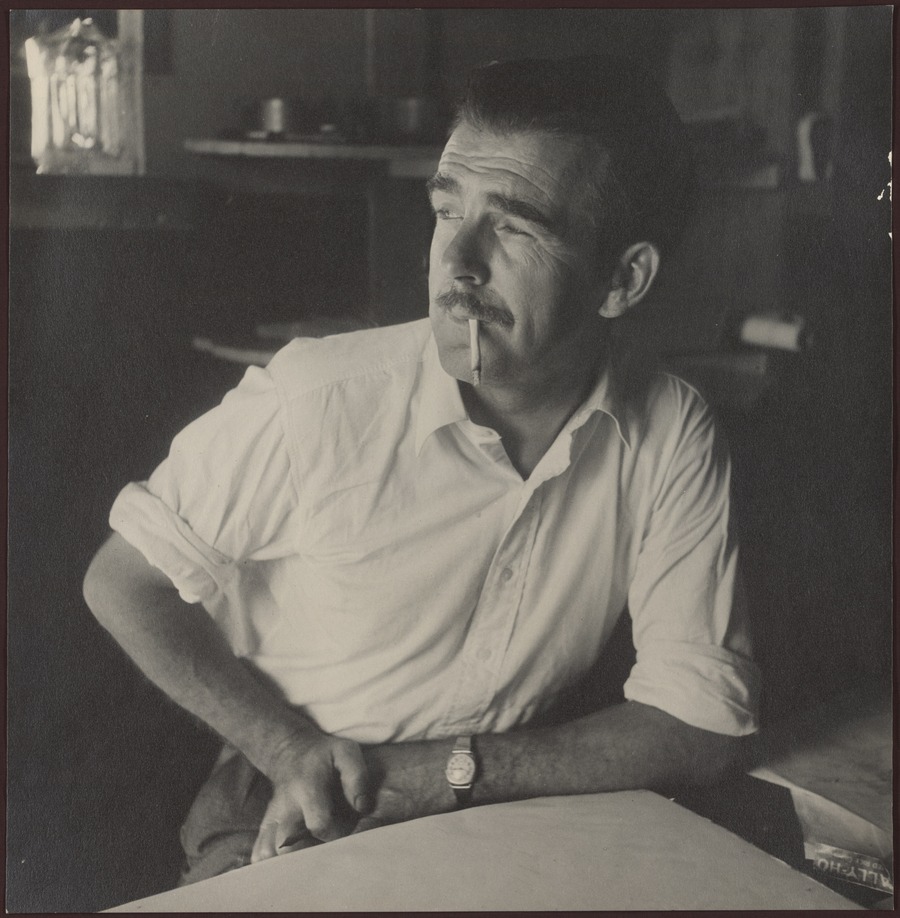 Black and white photograph of a man leaning against a table with a cigarette in his mouth