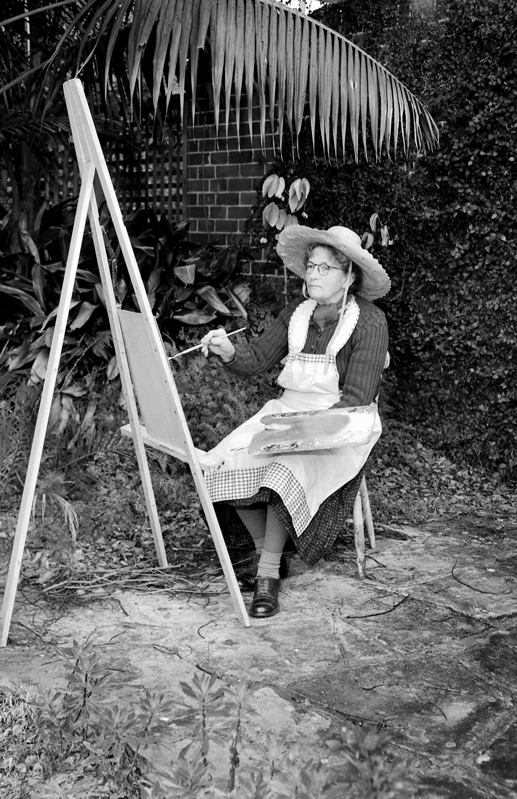 Image 4 of 6 - Black and white image of Grace Cossington Smith sitting outside as she paints. She is wearing an apron, glasses and a wide brimmed hat.