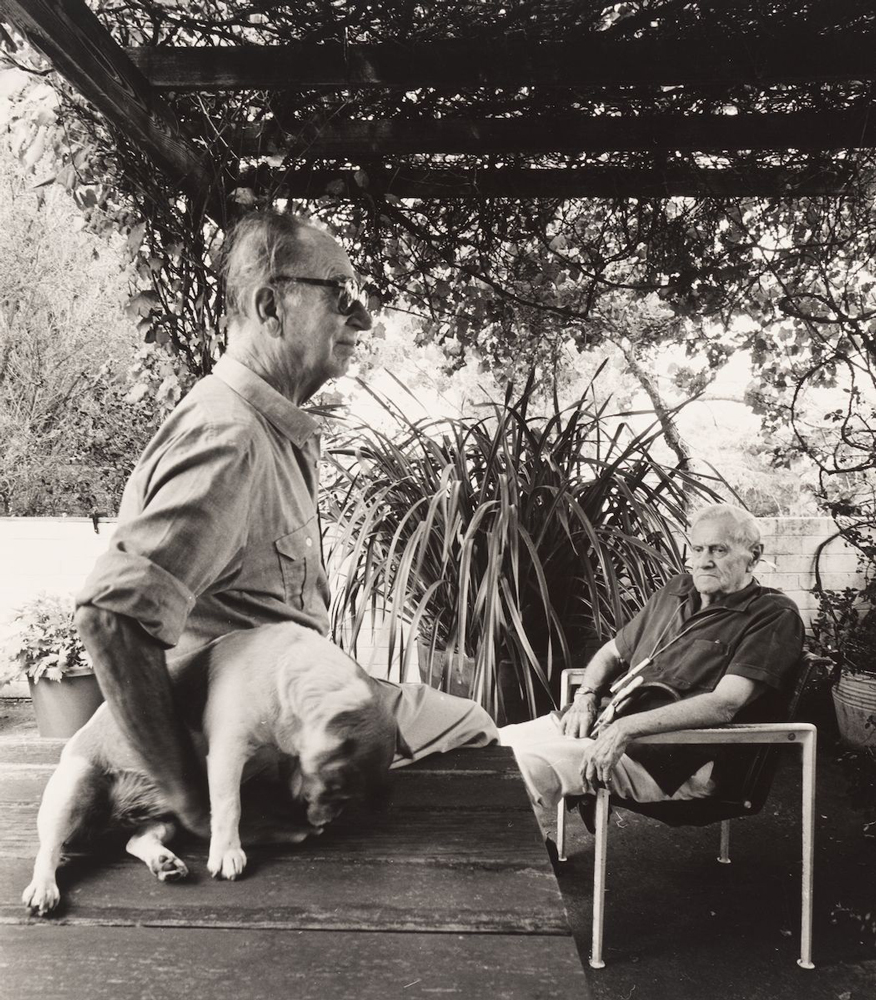 Black and white image of Patrick White sitting down and Manoly Lascaris holding a dog. Manoly is to the left of the image and is facing the right, he is wearing a button up shirt with the sleeves rolled up and has spectacles on. He is sitting on a table in the foreground with the dog and Patrick White is sitting on a chair to the right, slightly further back. The men are outside with plants and trees behind them.