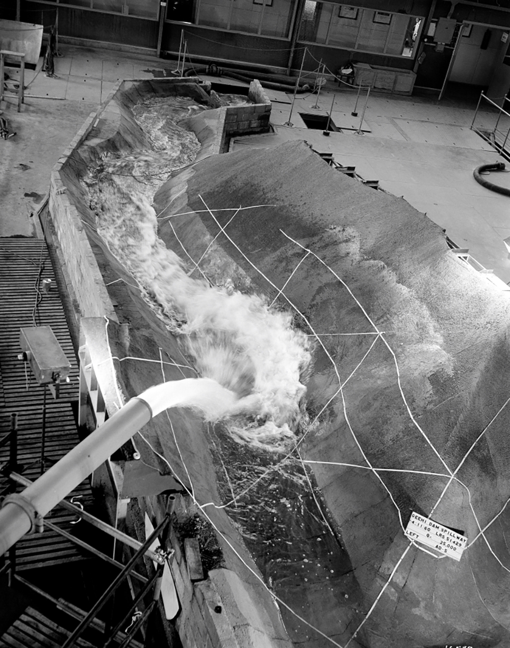 Image 3 of 5 - Black and white image of water gushing out of a pipe into a dam spillway model.