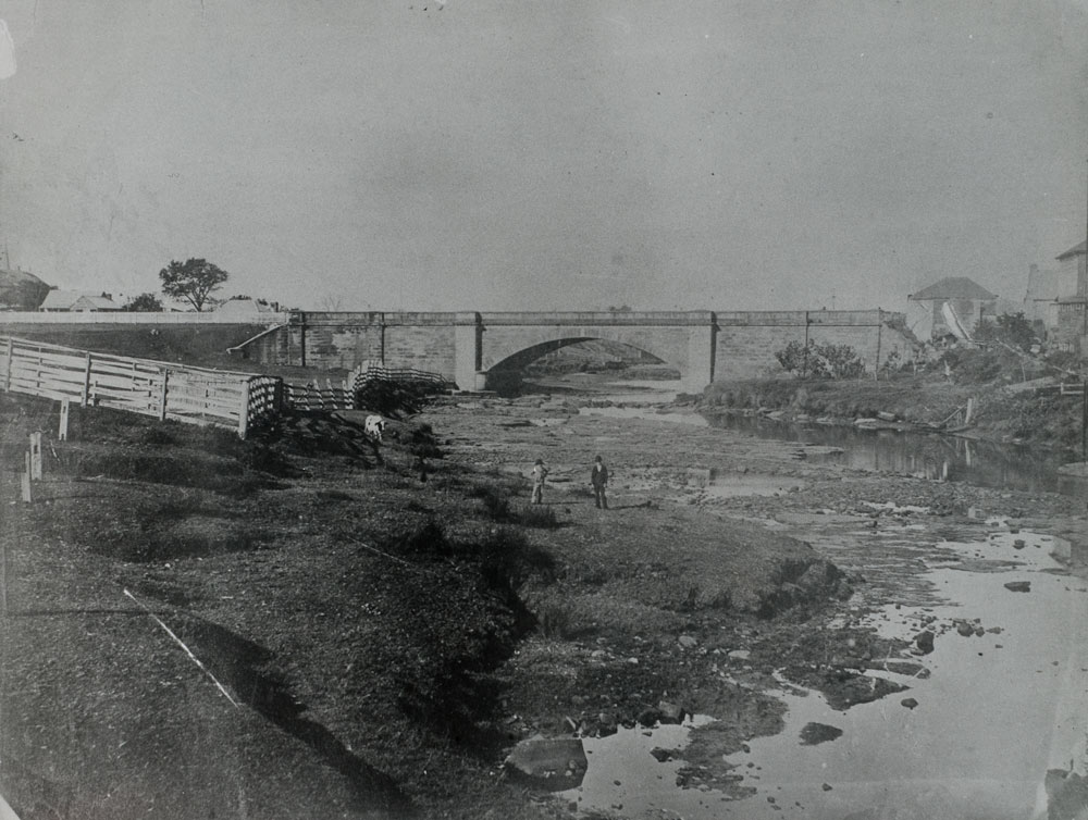 Image 2 of 3 - A photograph of the Lenox Bridge spanning the Parramatta River taken around 1880. Two gentleman and a cow are standing by the riverbank looking at the camera.